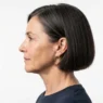 Side view of a 54-year-old woman with dark brunette blunt bob threaded with grey against a white backdrop