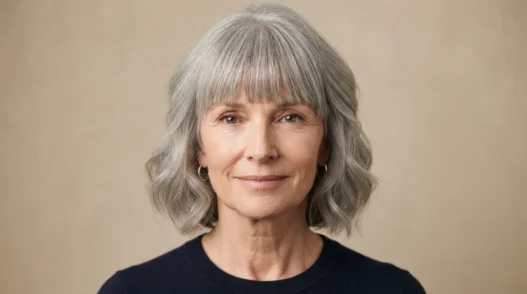 Woman in her late fifties with a long face wearing a full cheekbone-level fringe and soft grey waves at cheek level against warm beige backdrop