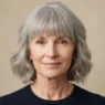 Woman in her late fifties with a long face wearing a full cheekbone-level fringe and soft grey waves at cheek level against warm beige backdrop