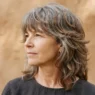 55-year-old woman with greying auburn modern shag, rough-dried to natural fall against warm clay backdrop in side light