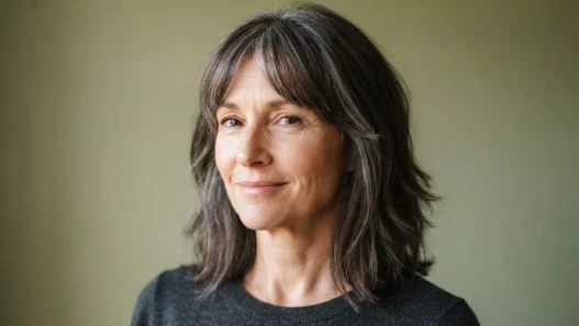 Woman in her early fifties with a mid-length lob and feathered side-swept bangs blending into layers against a sage backdrop