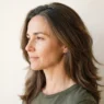 Side view of a 44-year-old woman with long layered brown hair showing first greys at the parting against a warm white backdrop