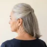 Rear three-quarter view of a 58-year-old woman with mid-length silver hair pinned half up at the crown against an ivory backdrop