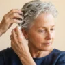 61-year-old woman with damp short silver hair applying mousse through mid-lengths with fingers against champagne backdrop