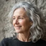 Woman in her late fifties with natural silver loose waves falling to the collarbone, soft window light on stone backdrop