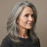 Woman aged 50 with thick greying hair in long interior-thinned layers against taupe backdrop, editorial lighting