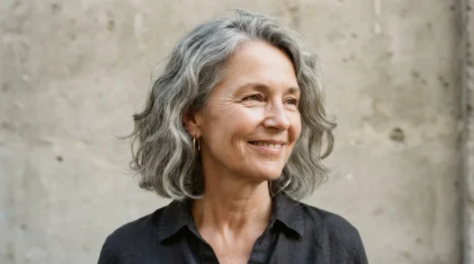 Sixty-year-old woman with square jaw and wavy grey lob against stone backdrop, wave sitting at jaw level