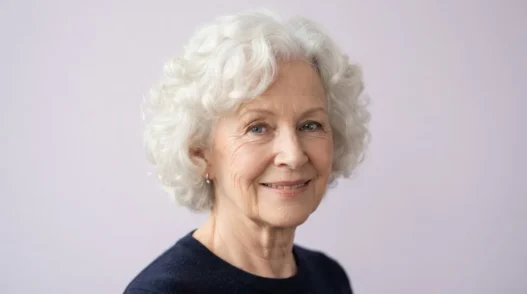 Elderly woman with soft white curls in a rounded set against a pale lilac backdrop, framed from crown to collarbone