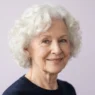 Elderly woman with soft white curls in a rounded set against a pale lilac backdrop, framed from crown to collarbone