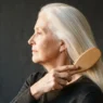 67-year-old woman with fine white hair being brushed smooth in profile view against charcoal backdrop with warm side light