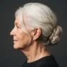 Profile view of older woman with white hair in a low chignon at the nape, warm side light on charcoal background