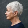 Side profile of a 66-year-old woman with a short white pixie cut against a plain slate backdrop with side lighting
