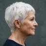 Profile view of a 66-year-old woman with a short white pixie cut, side light showing the taper at the nape and around the ear