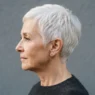 Profile view of a sixty-six-year-old woman with a short white pixie cut against a slate backdrop with side lighting on the nape taper